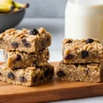 Freshly baked peanut butter banana oatmeal raisin bars stacked on a wooden board.